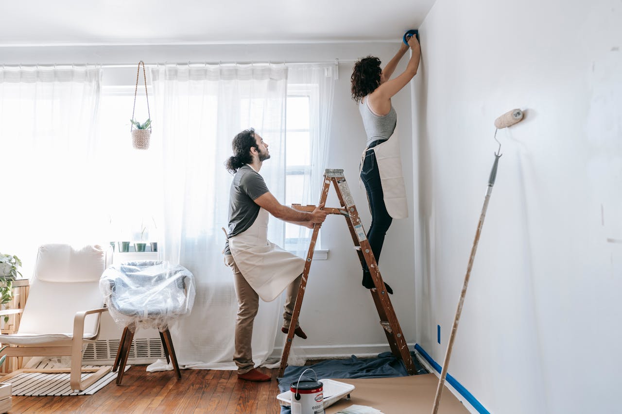 about-me Couple renovating their home, with man supporting ladder and woman working on walls indoors.